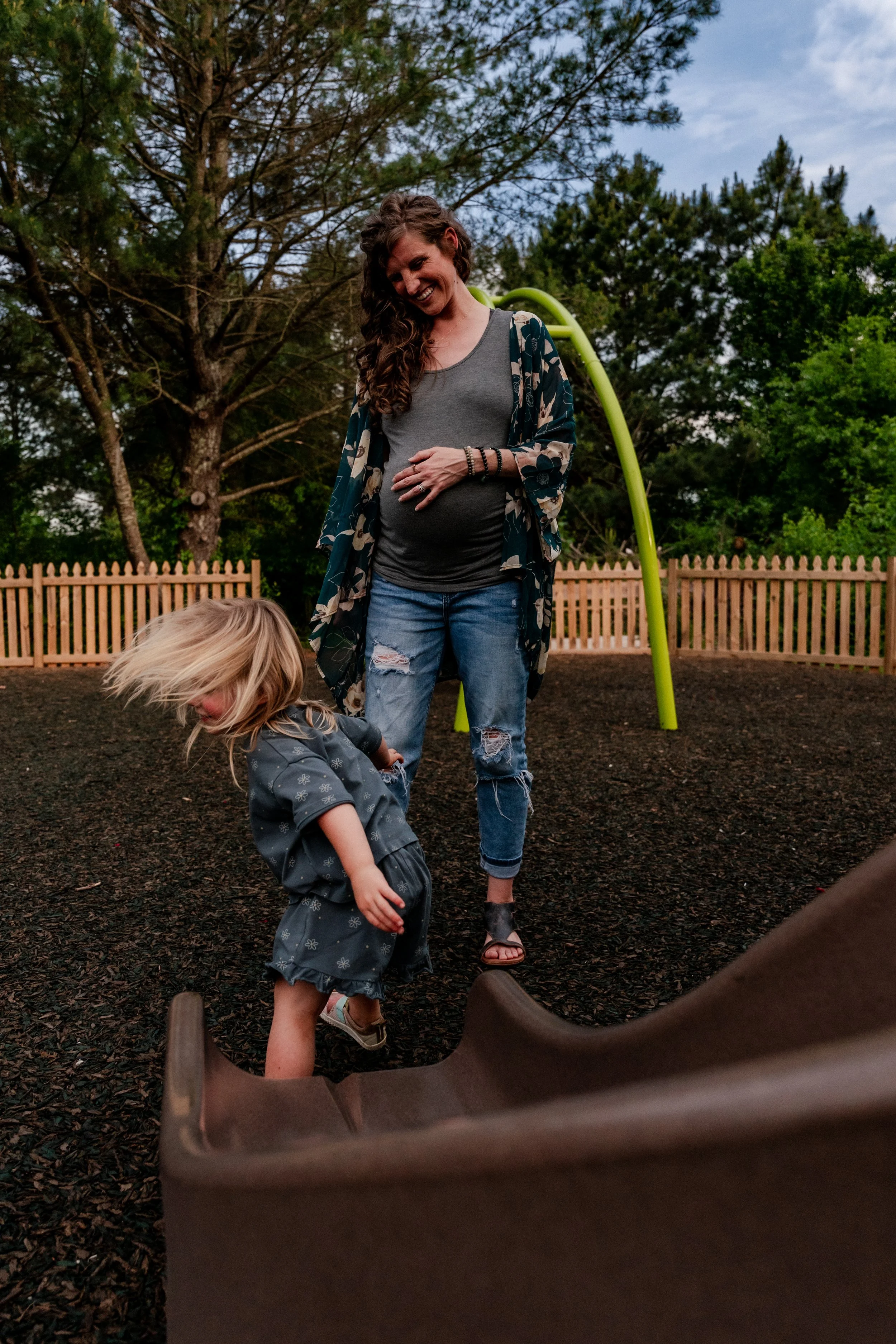 Pregnant mom laughing as her daughter comes down the slide at the playground