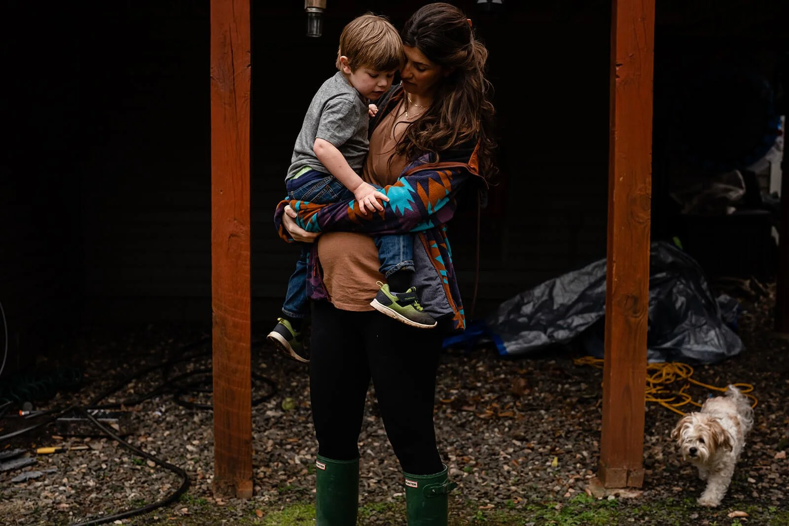 pregnant mom standing in her backyard while holding her toddler as the dog goes by