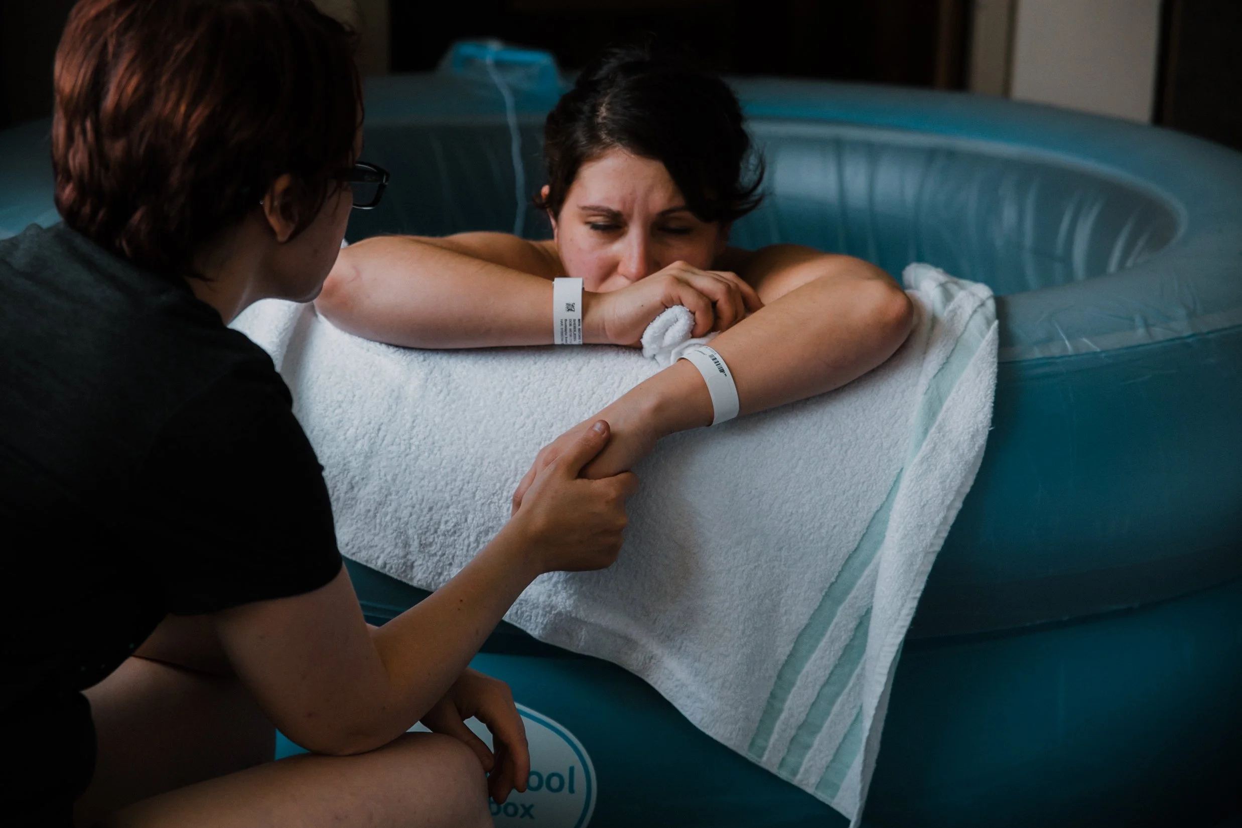 A woman in a birthing tub, in labor. The woman has hospital wristbands and is resting her head on her folded arms on a white towel. Her partner is gently holding her hand.