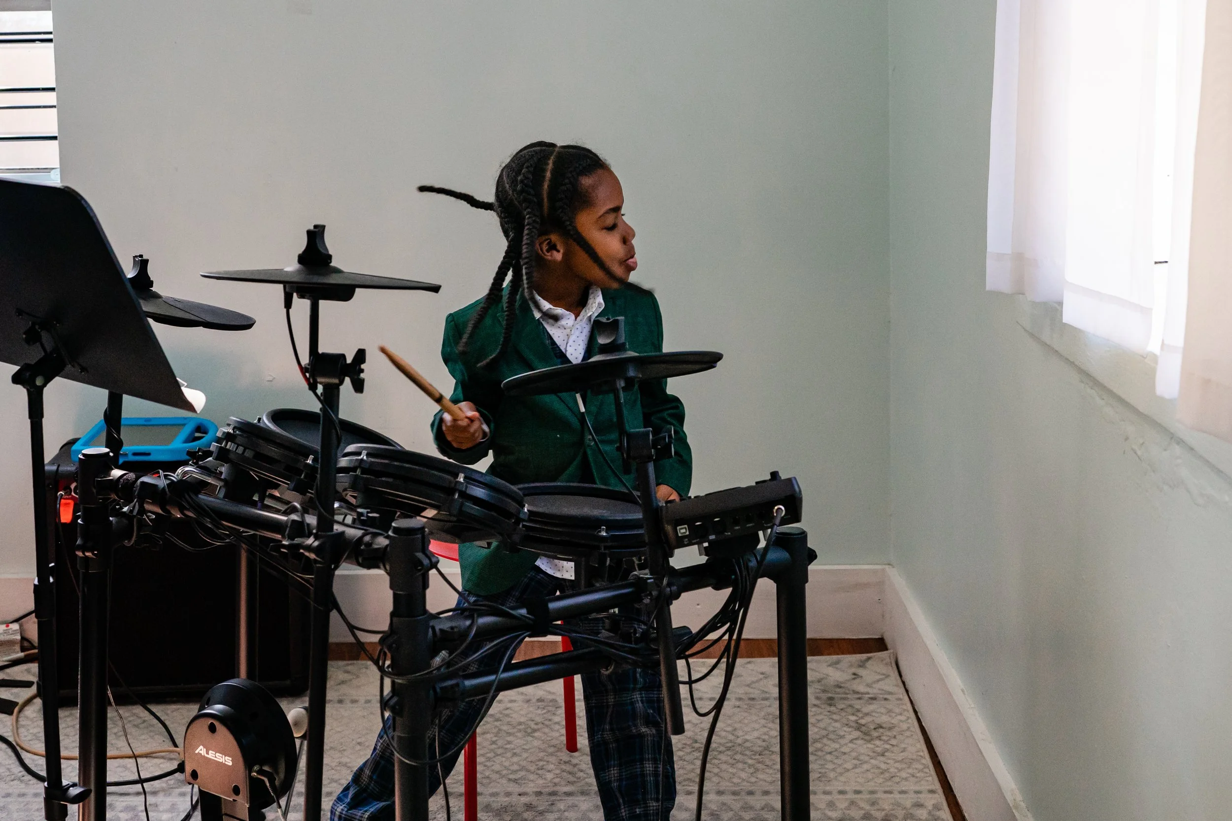 Candid family photography session in Atlanta with young boy playing an electronic drum set indoors, wearing a green blazer and checkered trousers, looking towards a window with white curtains.