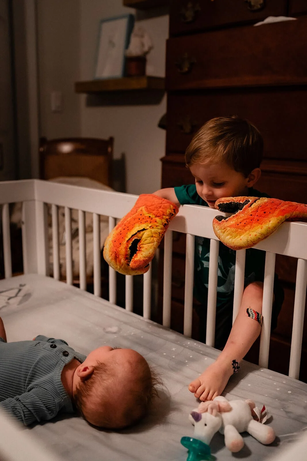 Big brother with oven mitts on, leaning over the edge of a crib to talk to his baby sister