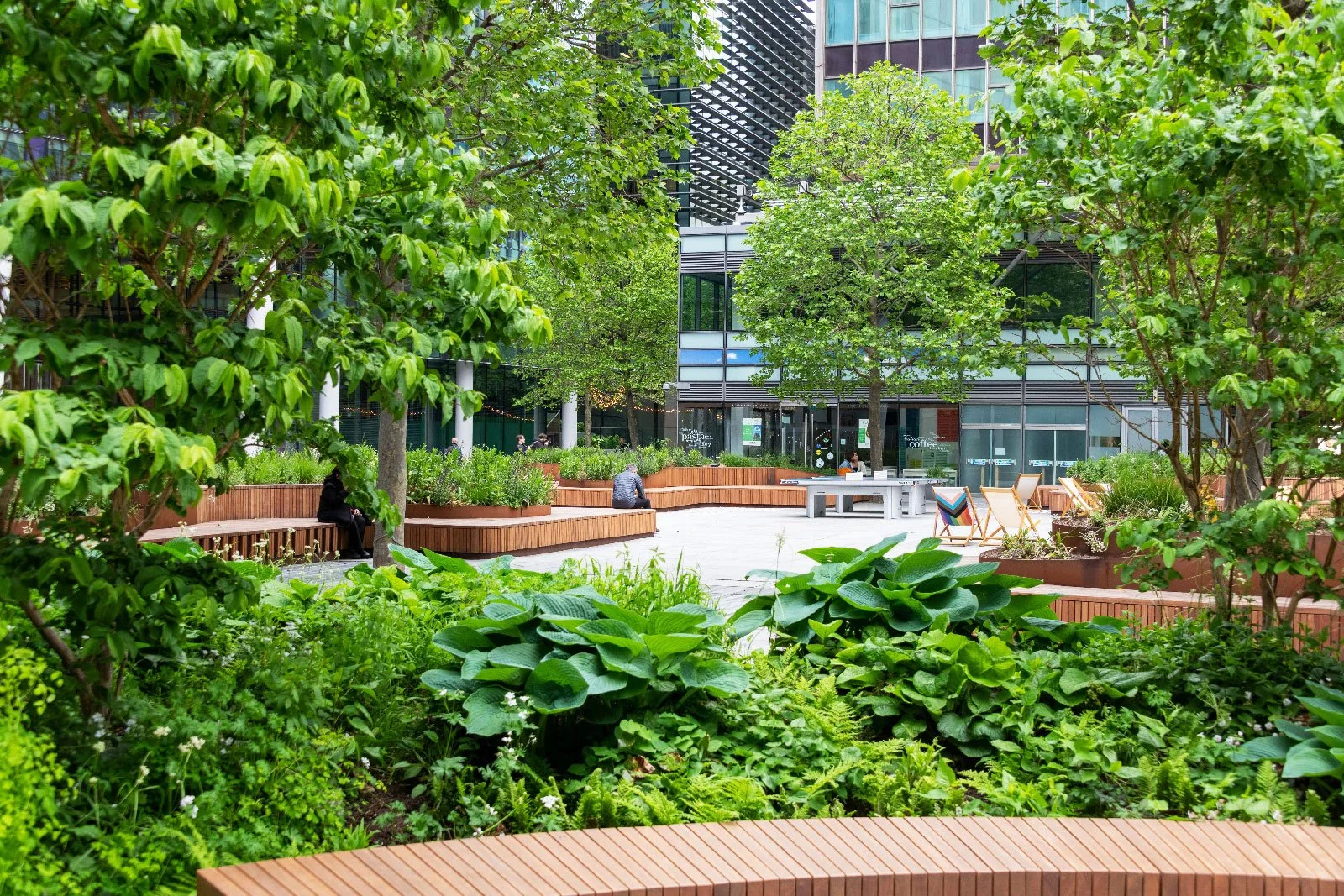 Urban park with lush green trees and plants, wooden benches, some people sitting and relaxing, and modern buildings in the background.