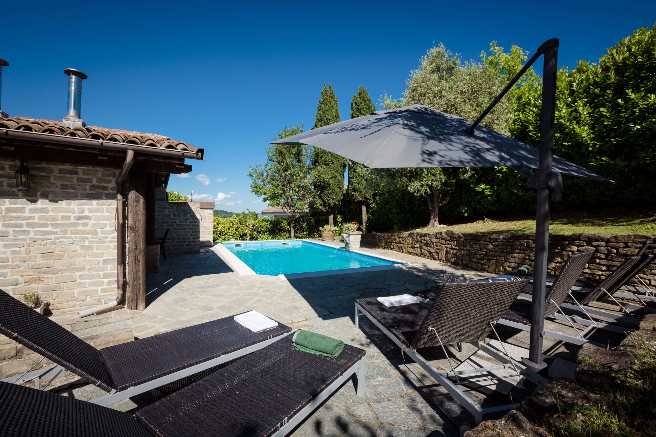 Outdoor swimming pool area with stone patio, lounge chairs, and umbrella next to a stone building, surrounded by greenery and trees, under a clear blue sky.