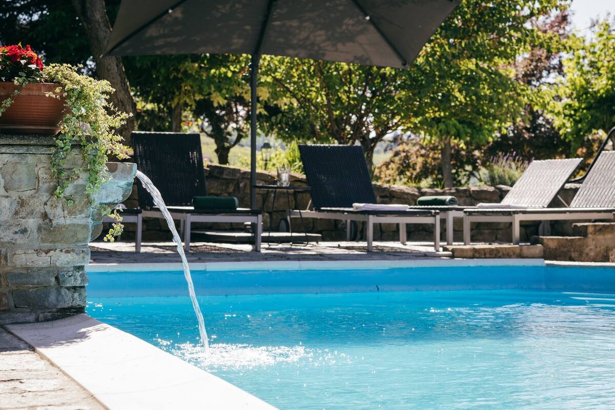 Swimming pool with flowing water, surrounded by stone patio, lounge chairs, and sun umbrella, with greenery in the background.