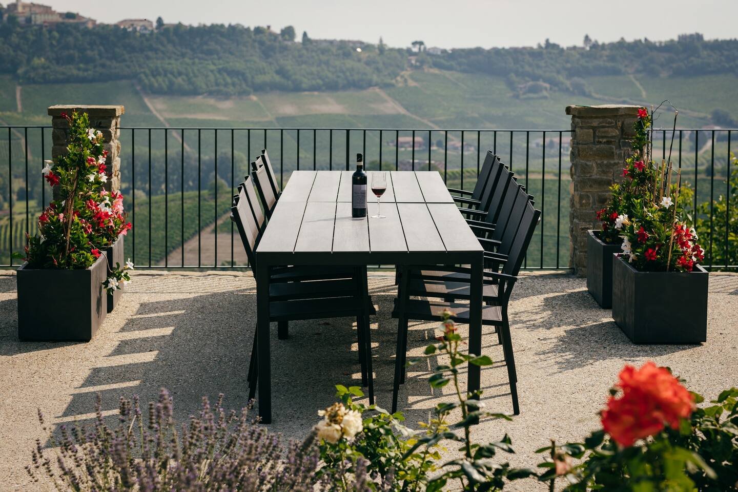 Outdoor patio with a black dining table and chairs, bottle of wine and glass, surrounded by flowers in planters, with a view of vineyards.
