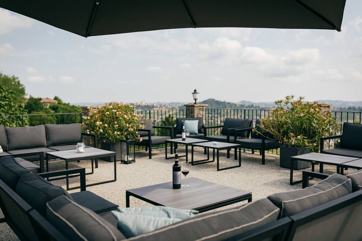 Outdoor patio with dark cushioned seating, tables, a bottle of wine, and potted plants, under a large umbrella, overlooking a scenic landscape.