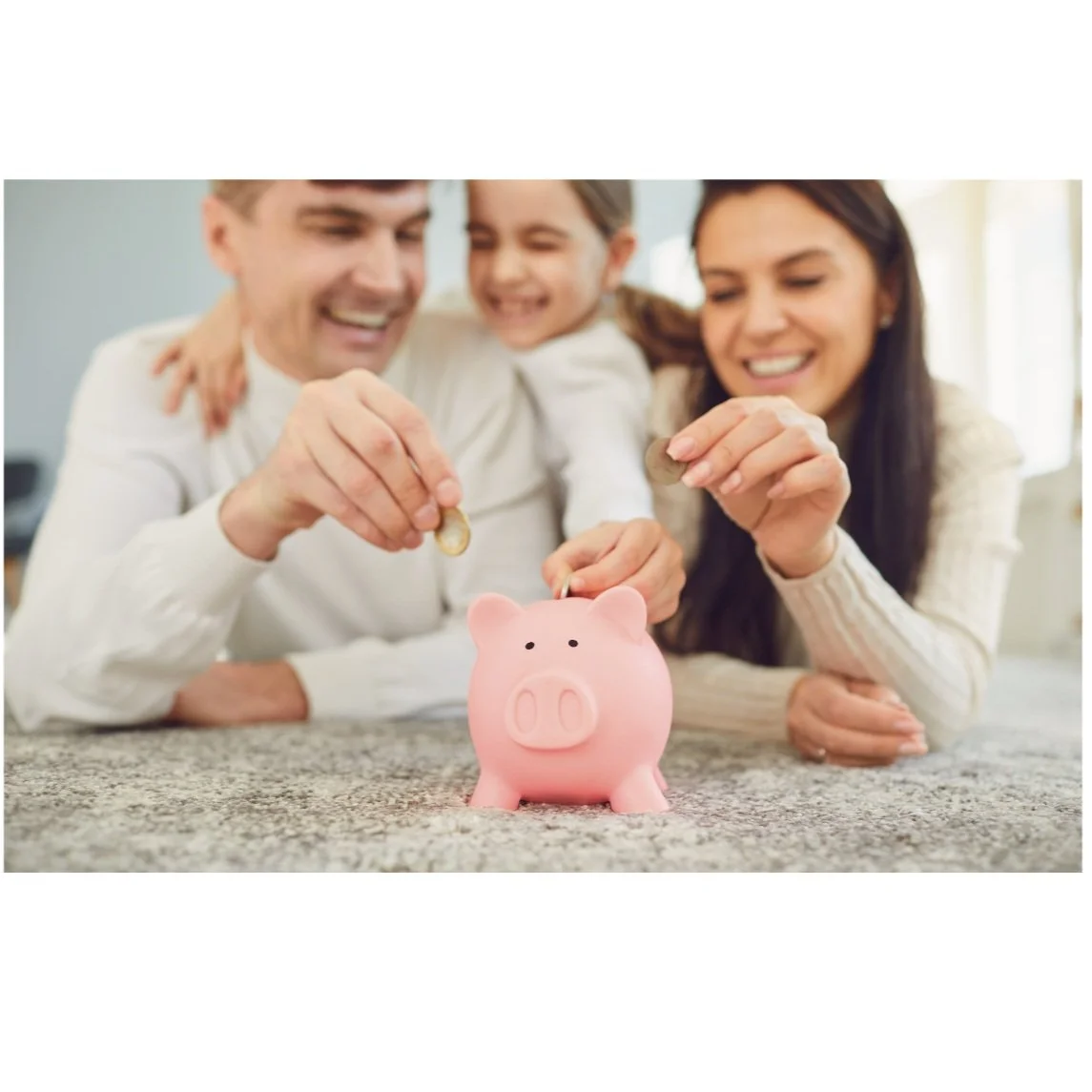 Family of three, smiling, putting coins into a pink piggy bank on a countertop.