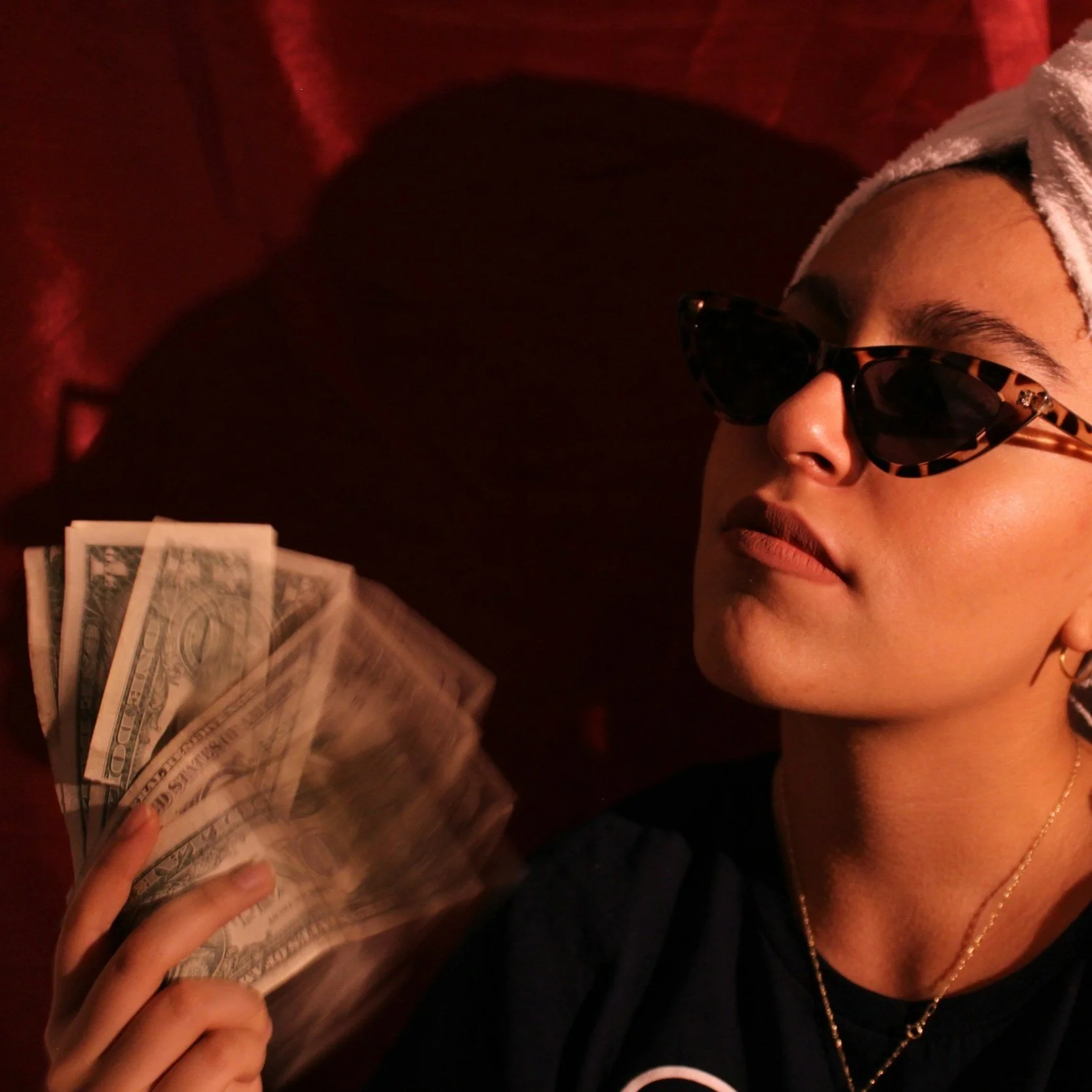 A person wearing sunglasses and a towel wrapped around their head is holding a fan of US hundred-dollar bills in a dark room with red lighting.