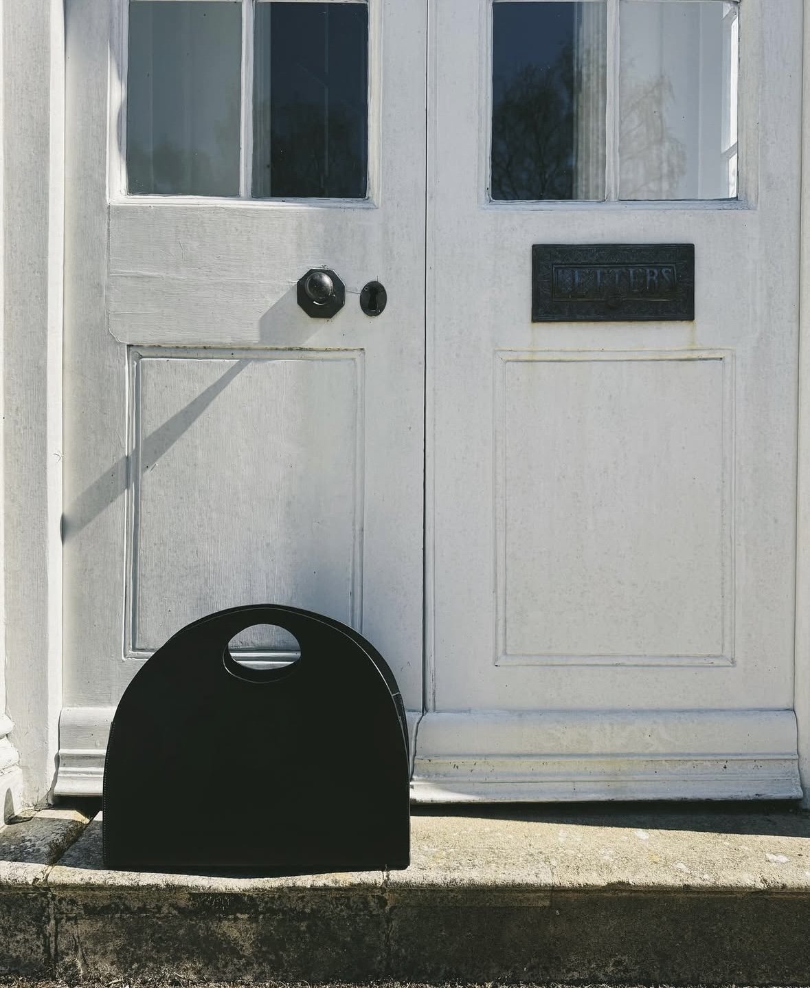 Large black statement tote bag with curved top and cut out handle on the step of a large house