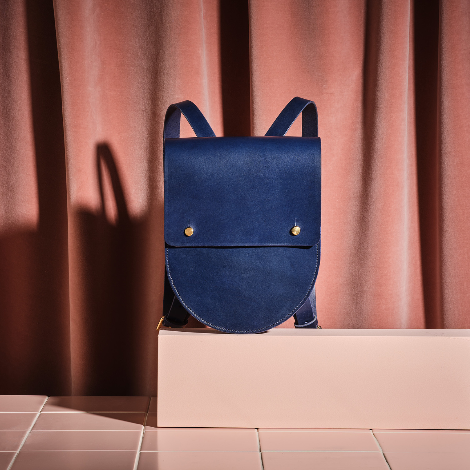 Navy leather rucksack with curved edge on a pink plinth and velvet curtain background