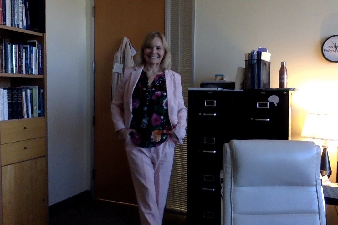 A woman standing in an office, smiling and wearing a pink suit and a floral blouse, with a bookshelf, a black filing cabinet, a white office chair, and a lamp in the background.