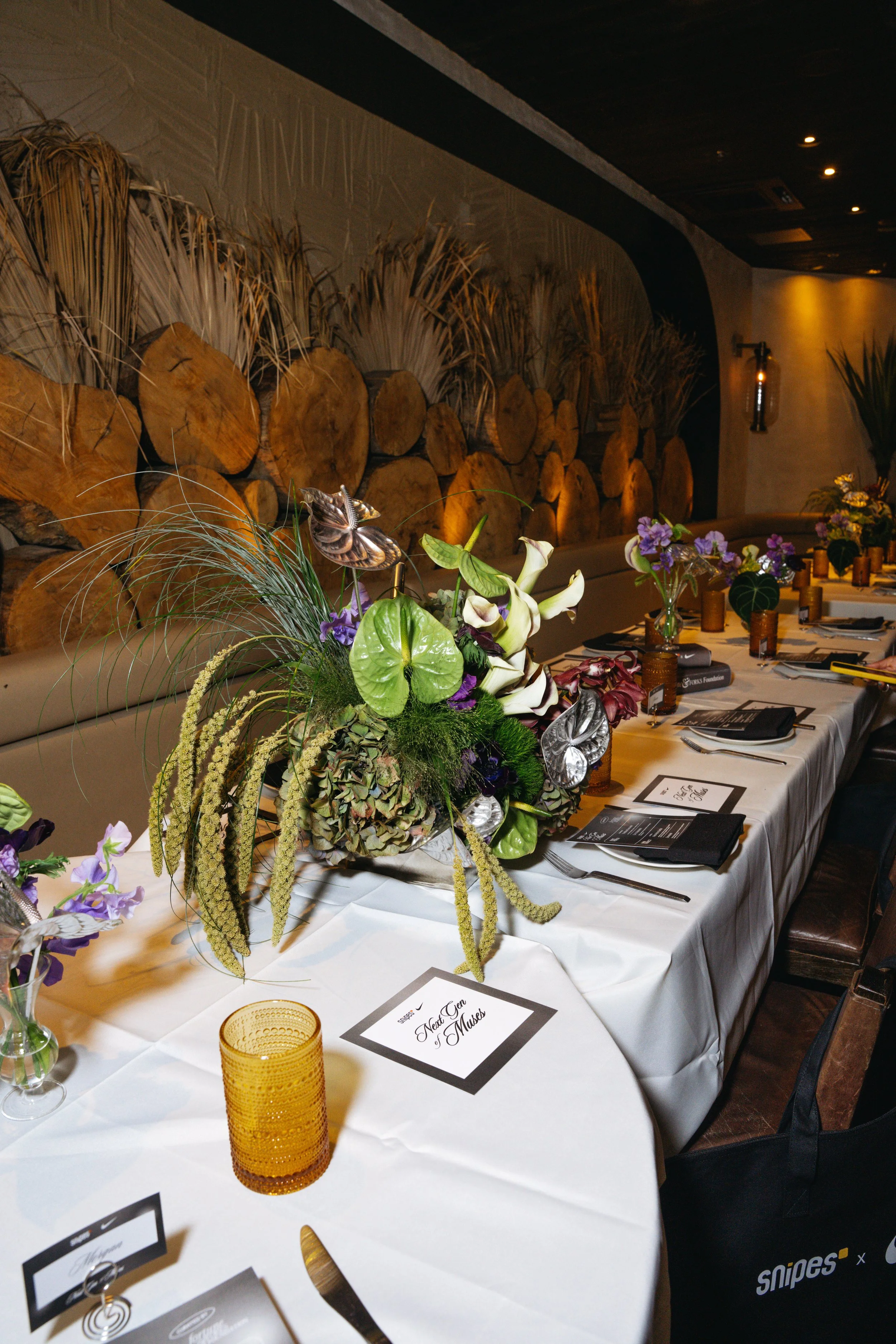 A long dining table decorated with floral arrangements, black napkins, and utensils, set for a formal event with a wood and stone wall background.