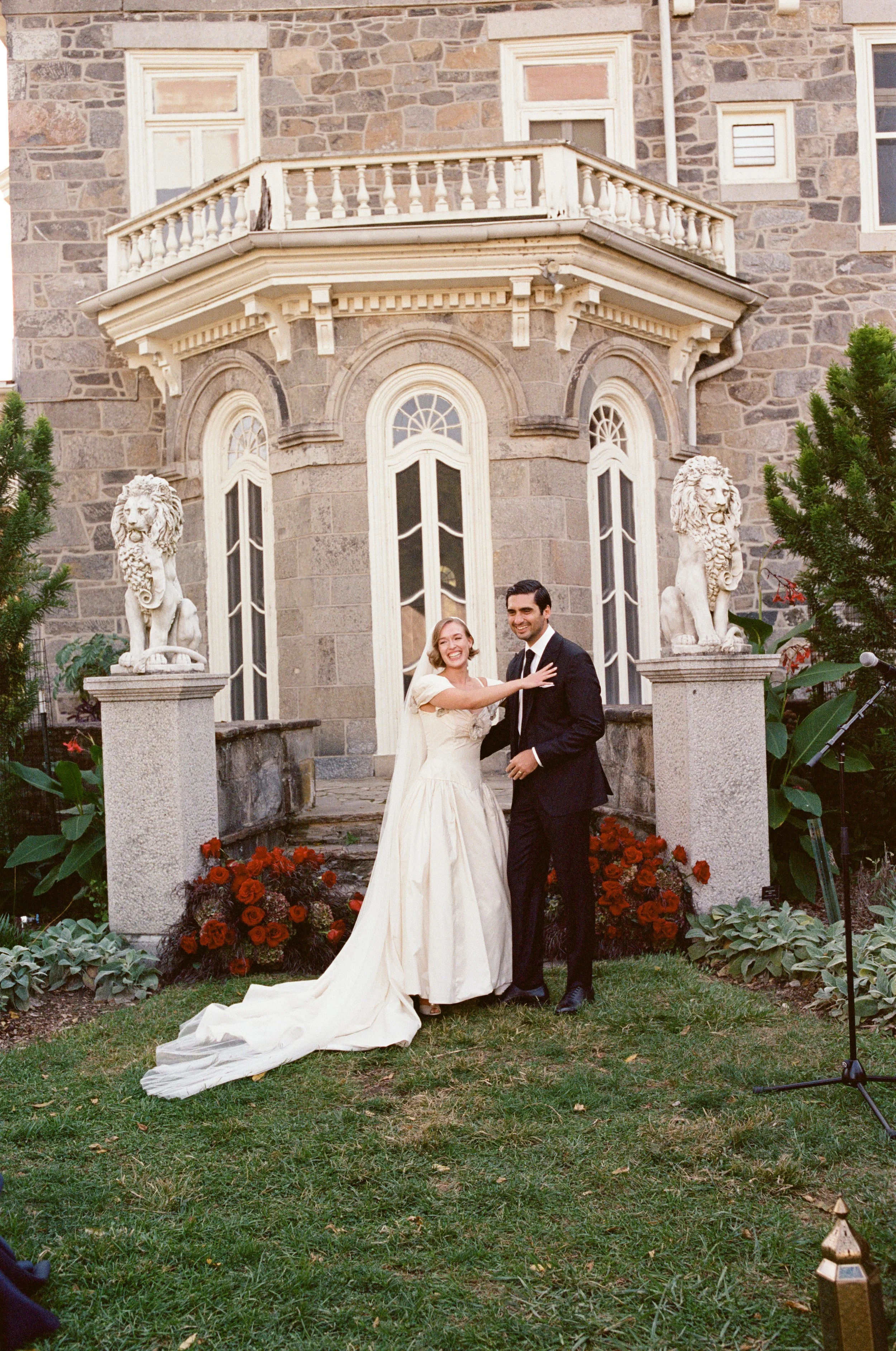 A bride and groom smiling and embracing in front of a stone mansion with ornate windows and lion statues, surrounded by garden flowers and greenery.
