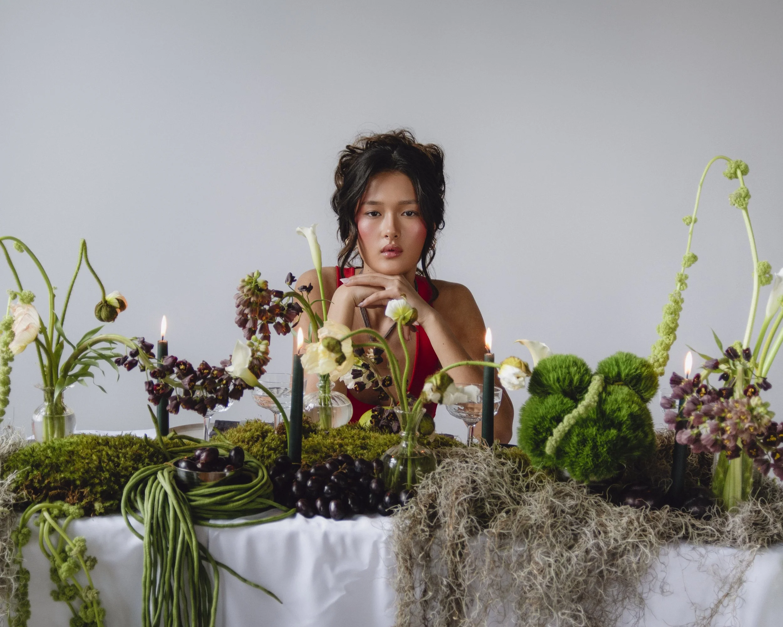 A woman in a red dress sitting behind a table decorated with flowers, moss, candles, and grapes, looking at the camera with her hands on her chin.