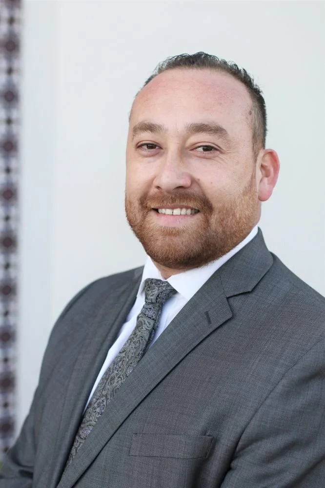 A man in a gray suit with a patterned tie, smiling, with a light-colored plain background.