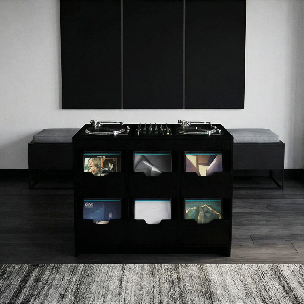 A modern black DJ station with turntables, vinyl records, and a gray cushioned bench on each side, set against a white wall with three black panels above, on a dark wooden floor with a patterned rug in front.