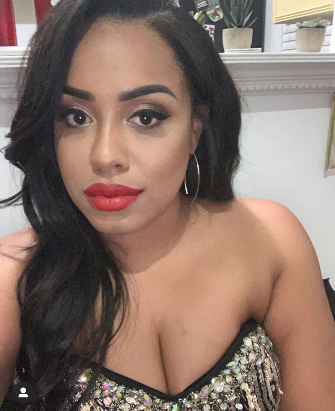 April Elizabeth Derokey A woman with long dark hair, wearing red lipstick and earrings, in a strapless dress decorated with sequins and beads, sitting indoors.