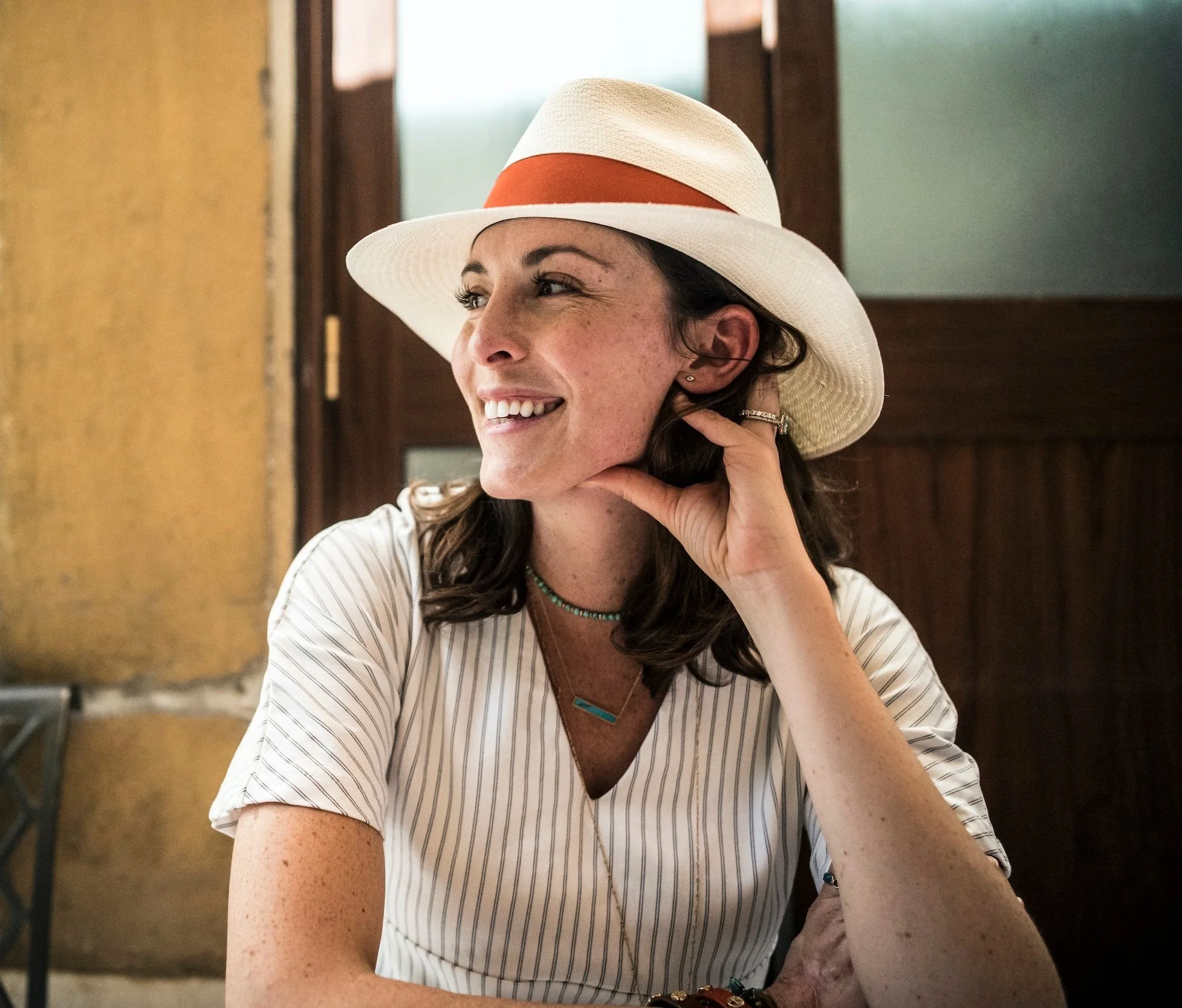 Victoria Fager A woman with dark hair, smiling, wearing a white straw hat with an orange band, sitting indoors near a window, resting her chin on her hand, dressed in a white shirt with thin stripes and layered necklaces.