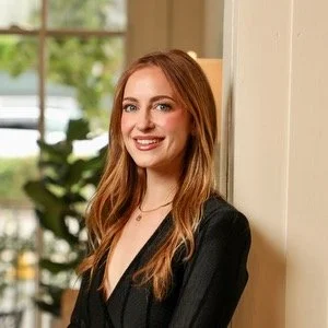 Julie Comarda Young woman with long red hair smiling, wearing a black blazer, standing indoors near a window with greenery outside.