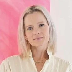 Kate Grace Bauer Rouchelle A woman with blonde hair and a neutral expression, wearing a white top, standing against a pink and white background.