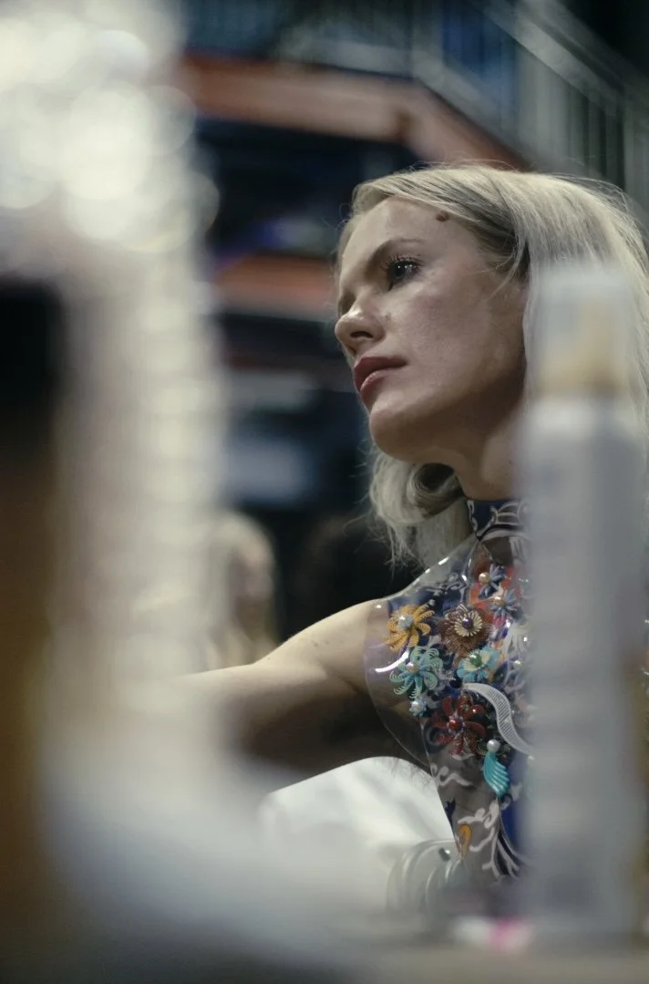 Brittany Schall L'adorna A woman with blonde hair and fair skin looks to the side, seated in a dimly lit indoor setting, surrounded by blurred objects.