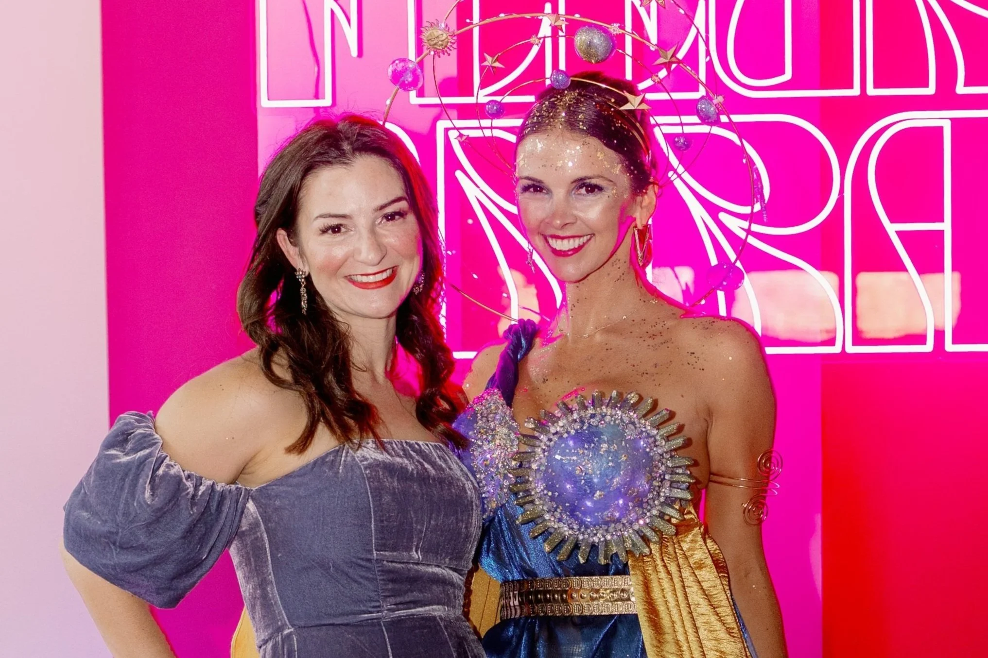 Two women smiling, standing in front of a bright pink neon sign background. One woman wears a purple velvet off-the-shoulder dress, and the other woman is dressed in a cosmic-themed costume with glitter, jewelry, and a star-themed headpiece.