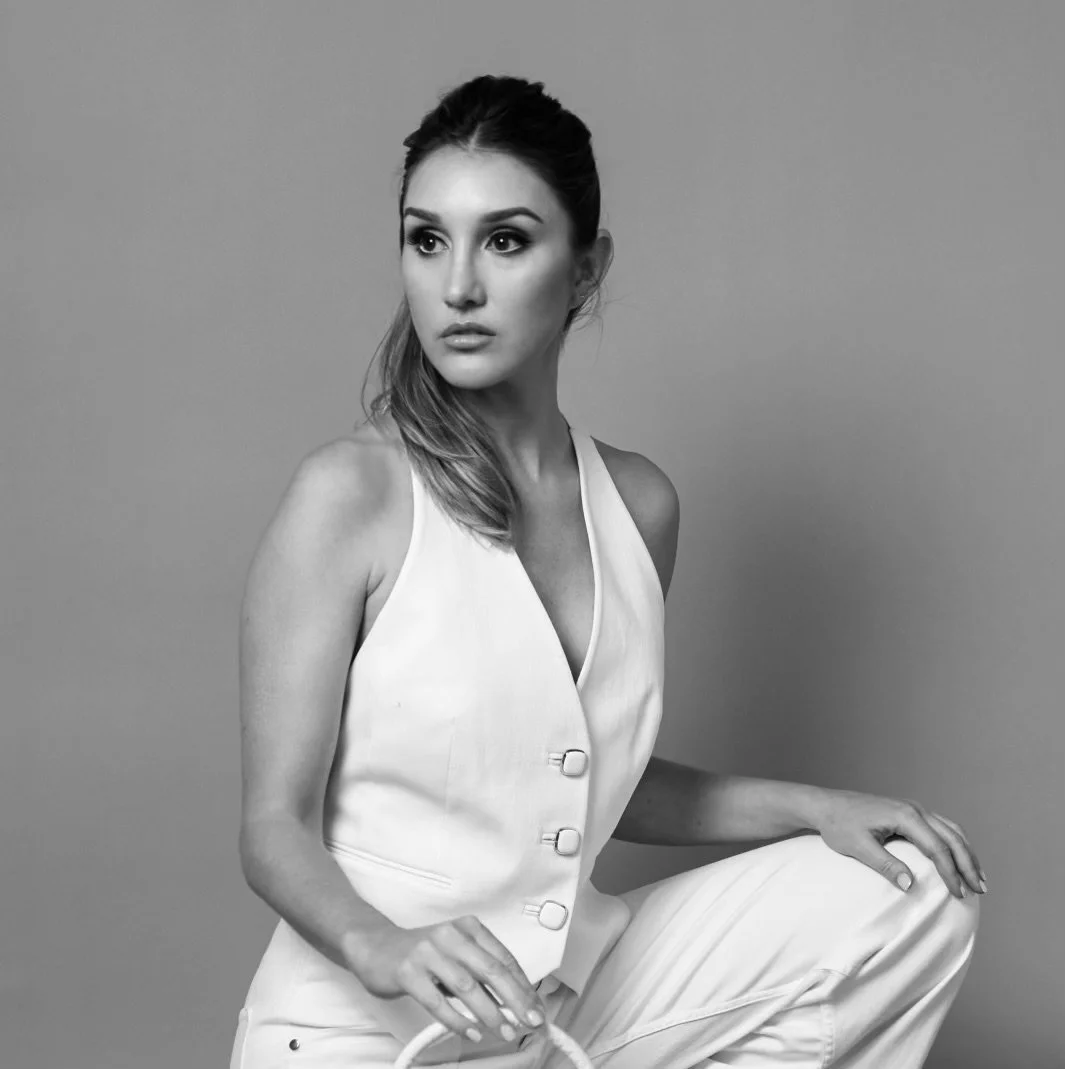 Cece Villere Black and white photo of a woman with long hair, wearing a sleeveless white vest and pants, sitting with one knee up, against a plain background.