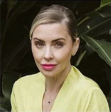 Melissa coleman A woman with blonde hair wearing a light yellow top, posing outdoors with green foliage background.