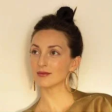 A woman with dark hair in a top bun, wearing gold hoop earrings and a beige top, against a neutral background.