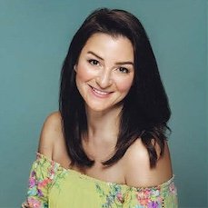 Jackie Haxthausen A woman with long dark hair smiling, wearing an off-shoulder floral top against a teal background.