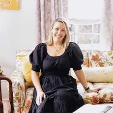 Woman sitting on a floral sofa, smiling, in a room with large windows and curtains.