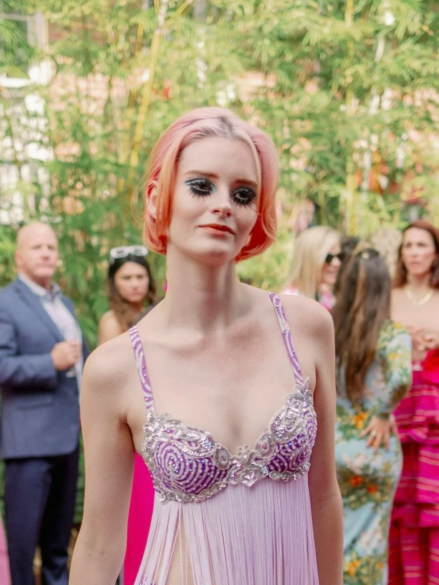 A woman with short pink hair wearing a purple and pink dress with sequined details, standing outdoors with a crowd of people in the background.