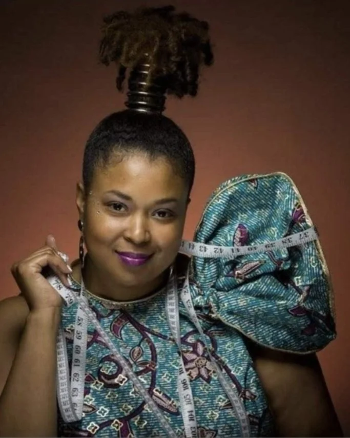 Renee Johnson Woman with natural hair styled in a high puff, wearing traditional African clothing, holding a measuring tape, against a brown background.