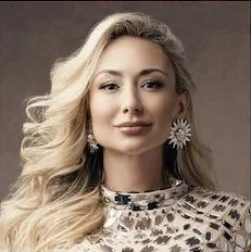 Reagan Charleston
Thomas, Esq Portrait of a woman with long blonde curly hair, wearing floral earrings and a patterned top, against a neutral background.