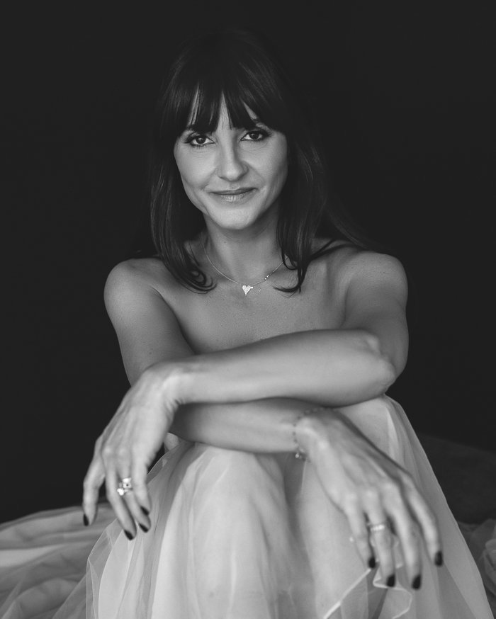 Shelley Dee-Girgrah A black and white portrait of a woman with shoulder-length dark hair and bangs, smiling at the camera, wearing a necklace with a heart pendant, and a light, flowing dress, sitting with her arms resting on her knees