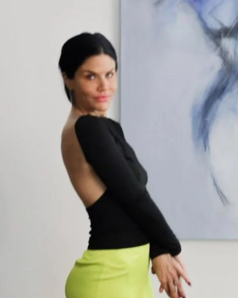 Brittany Lee Howard Woman with black hair, wearing a black top and yellow skirt, standing indoors beside an abstract painting.