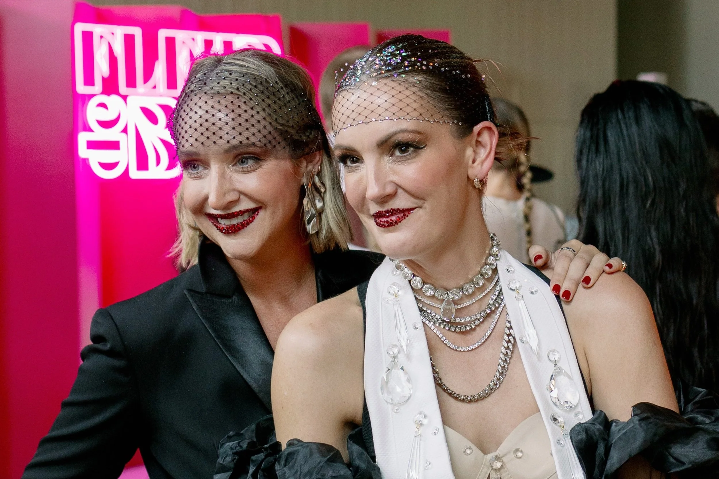 Two women dressed in glamorous outfits with glitter and jewelry smiling at an event with a pink sign in the background.