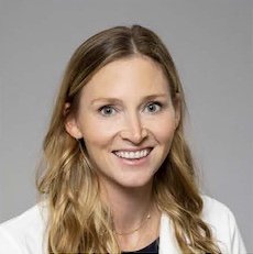 Dr. Caitlin Taylor A smiling woman with long, wavy blonde hair wearing a white blazer over a dark top, posed against a neutral gray background.