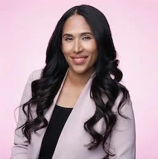 A woman with long, wavy black hair smiling against a light pink background, wearing a light-colored blazer over a black top.