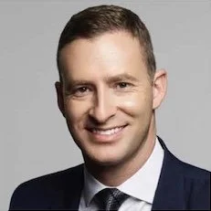Jonathan Bank A man in a suit smiling against a gray background.