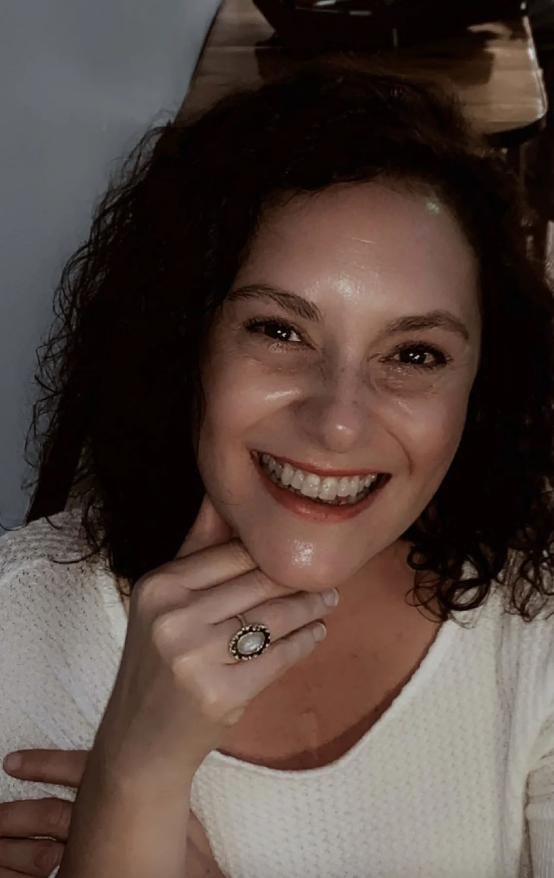 Kristen Monroe A woman with curly dark hair smiling, showing her teeth, wearing a white top and a ring on her finger, sitting indoors.