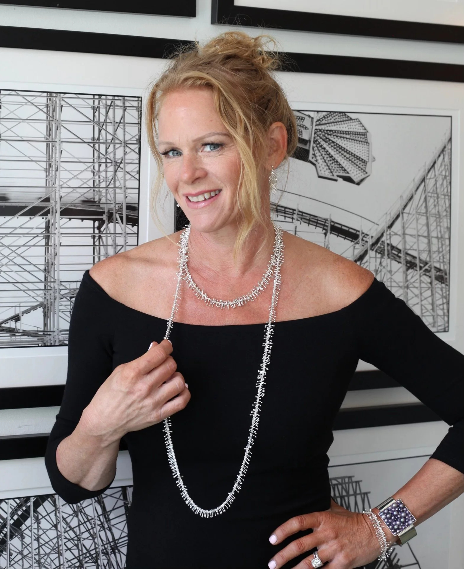 Dominique Giordano A woman with blond hair styled in a messy bun, wearing a black off-the-shoulder top and silver jewelry, standing in front of black and white architectural posters.