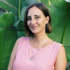 A woman with shoulder-length brown hair, wearing a pink sleeveless top and a necklace, standing outdoors in front of green plants.
