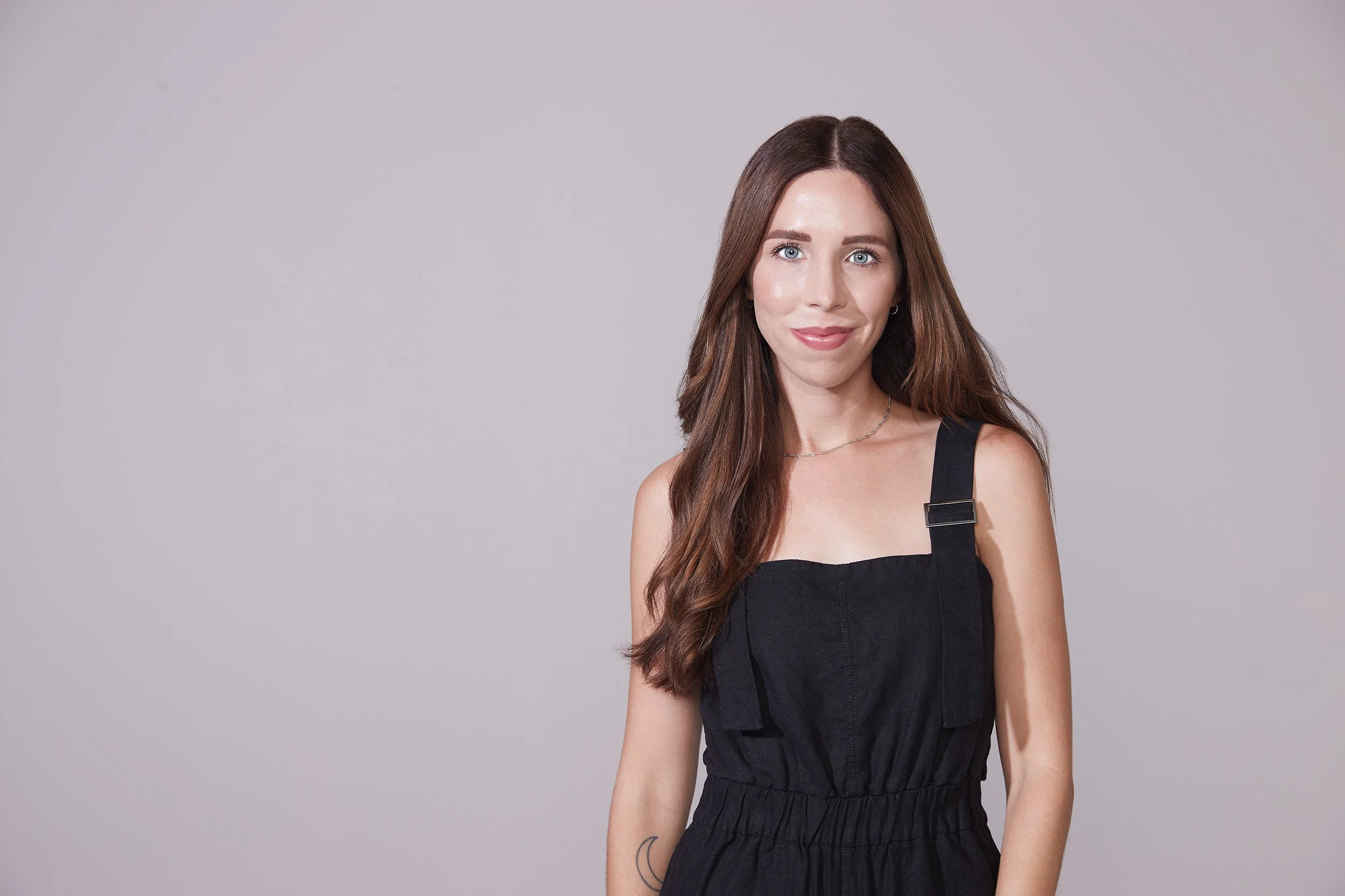 Janna Hart Black A young woman with long brown hair, blue eyes, and fair skin standing against a light gray background, wearing a black sleeveless jumpsuit with straps and smiling at the camera.