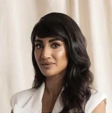 A woman with dark hair styled in loose waves, wearing a white blazer, standing in front of light-colored curtains.
