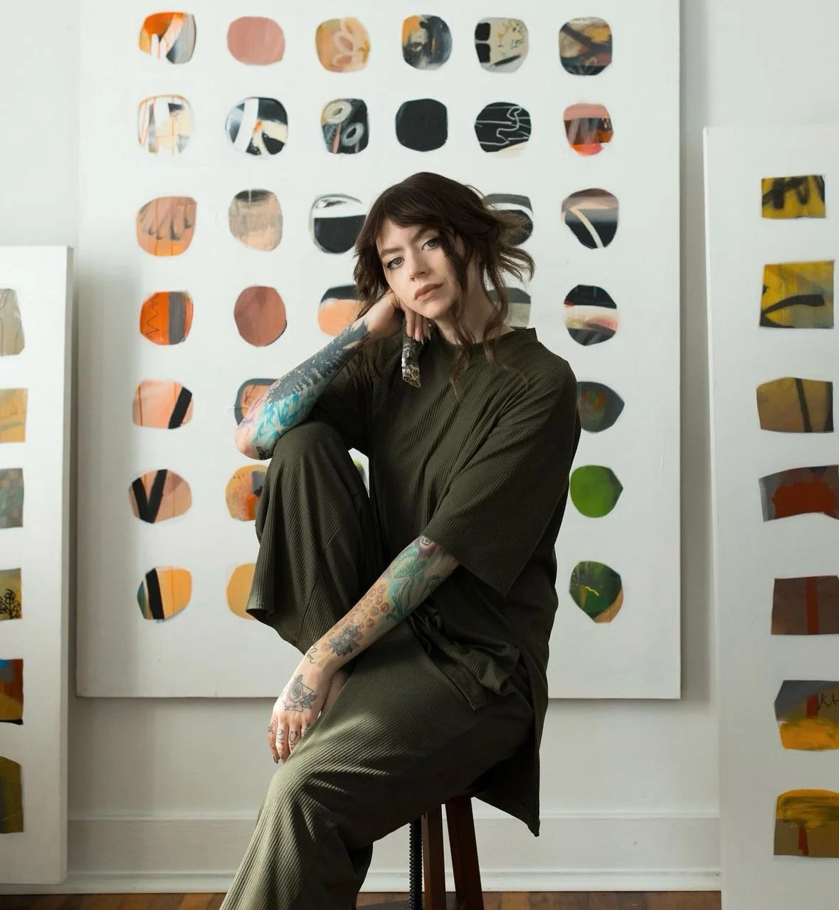 Kim Meadowlark A woman with dark hair and tattoos sitting on a stool in front of colorful, abstract artwork on a white wall.