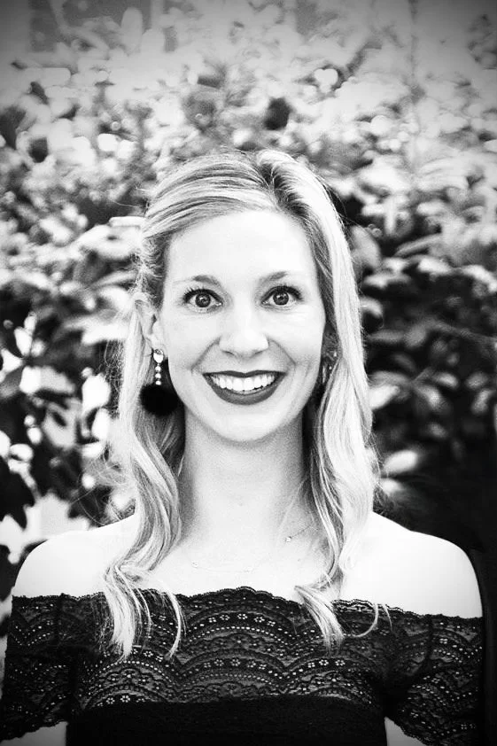 Caroline Jacobson A smiling woman with shoulder-length wavy hair, wearing a lace off-the-shoulder top and earrings, standing in front of a bushy background.