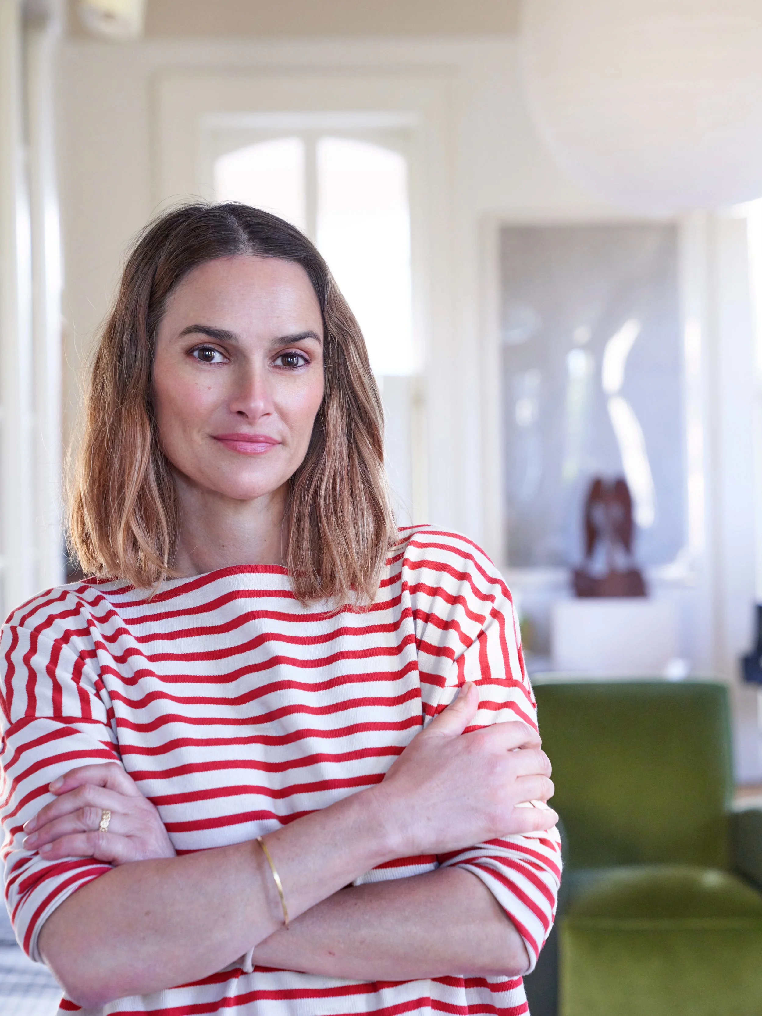 Sara Costello A woman with shoulder-length brown hair, wearing a red and white striped shirt, standing indoors with arms crossed, looking at the camera.