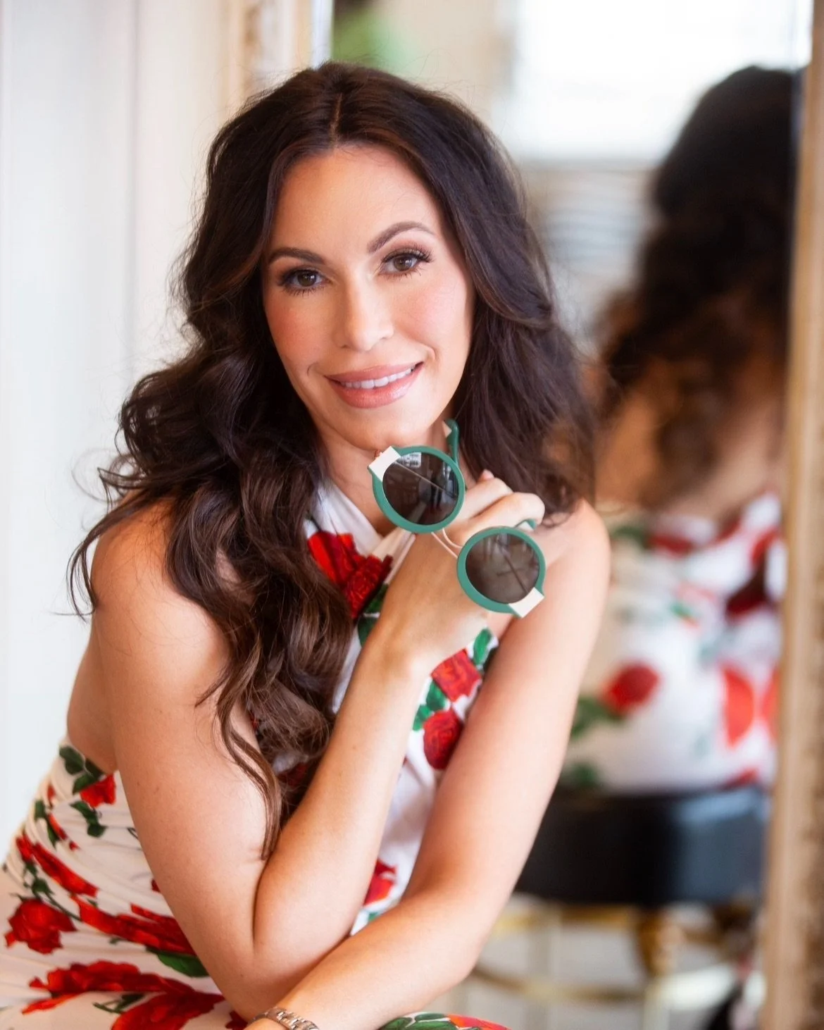 Lauren Agnew A woman with long, dark, wavy hair and light skin, smiling and holding a pair of sunglasses, standing in front of a mirror, wearing a floral dress with red and green pattern.