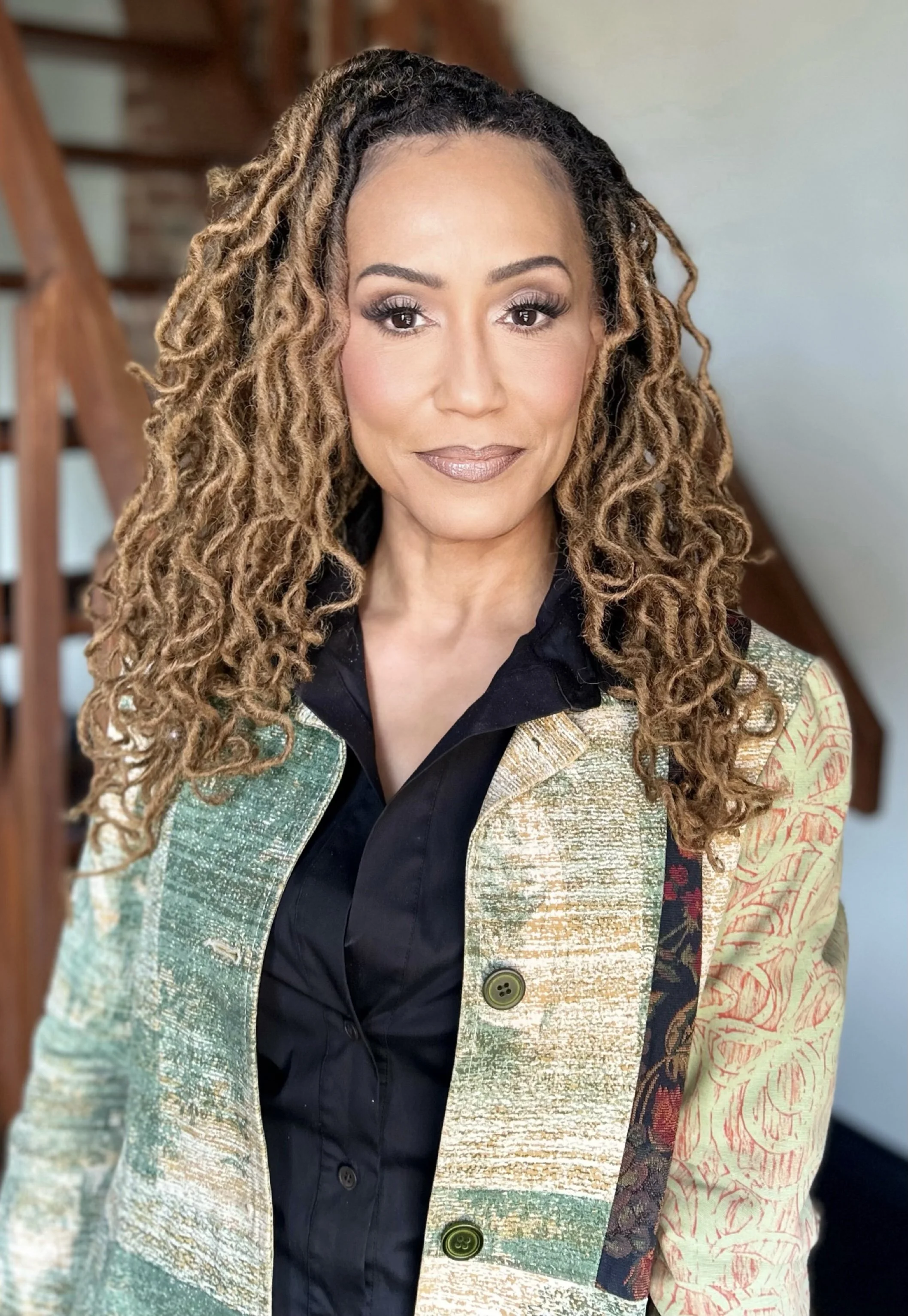 Tracee Dundas A woman with long, curly, blonde-brown hair, wearing a colorful, patterned jacket over a black shirt, standing indoors.